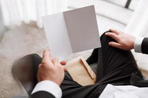 the groom holds an envelope with a letter from the bride. man is reading and sitting in the chair near the window. A letter for the groom. declaration of love. mock up place photo