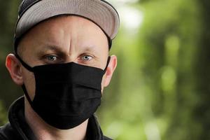 Young man in black protective antiviral mask in the summer park. The guy is resting outdoors on a sunny summer day in a mask made by his own hands. Virus protection. Quarantine measures photo
