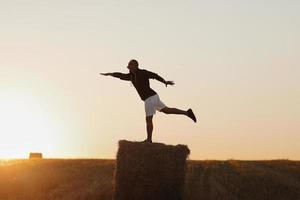 handsome young man in black sweatshirt and white short, in summer field outdoor, having fun on hay bale, haystack on sunset photo