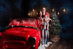 Young beautiful parents holding their little cute daughter in their arms having fun near retro car in studio. Christmas family look. New Year's scenery. photo