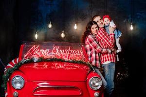 jóvenes y hermosos padres sosteniendo a su pequeña y linda hija en sus brazos divirtiéndose cerca de un auto retro en el estudio. aspecto familiar navideño. paisaje de año nuevo. foto