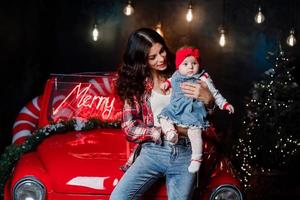 una mujer feliz con una niña con un lazo rojo en la cabeza está sentada y divirtiéndose en un auto retro en el estudio de navidad. mamá e hija están jugando adentro. foto