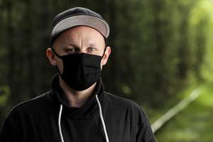 Young man in black protective antiviral mask in the summer park. The guy is resting outdoors on a sunny summer day in a mask made by his own hands. Virus protection. Quarantine measures photo