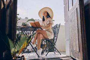 Beautiful young woman holding book and looking happy while relaxing on the balcony photo