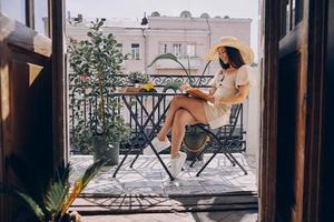 Happy young woman in elegant hat reading book while relaxing on the balcony photo