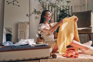 Attractive young woman packing clothes to suitcase and smiling while sitting on the floor at home photo