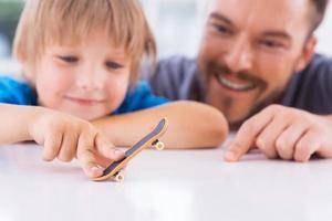 How are you doing like this Cheerful father looking at his sun playing with fingerboard and smiling photo