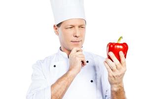 eligiendo las mejores verduras para su comida. alegre chef maduro con uniforme blanco sosteniendo pimiento rojo y mirándolo mientras se enfrenta a fondo blanco foto