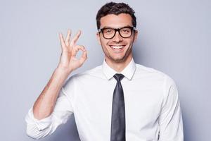 todo está bien, un joven feliz con camisa y corbata haciendo un gesto de ok y sonriendo mientras se enfrenta a un fondo gris foto