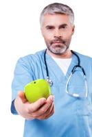 Join healthy lifestyle Confident mature surgeon in blue uniform stretching out a green apple while standing isolated on white photo