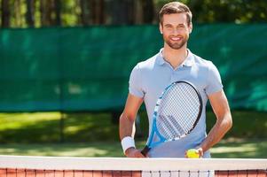 Great day to play Cheerful young man in polo shirt holding tennis racket and ball while standing on tennis court photo