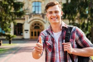 pulgar hacia arriba para el éxito estudiante guapo mostrando su pulgar hacia arriba y sonriendo mientras está de pie al aire libre con el edificio de la universidad en el fondo foto