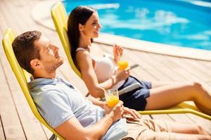 What a great day Side view of beautiful young couple holding cocktails and smiling while sitting at the deck chairs by the pool photo
