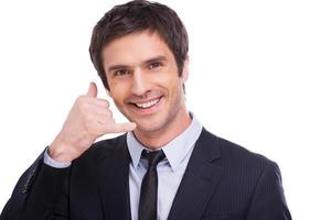 Llámame joven feliz con camisa y corbata haciendo gestos con el teléfono móvil cerca de su cara y sonriendo mientras está aislado en el fondo blanco foto