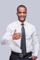 pulgar hacia arriba para el éxito confiado joven africano con camisa y corbata mostrando su pulgar hacia arriba y sonriendo mientras está de pie contra un fondo gris foto