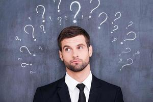 So many questions Confused young man standing against blackboard with question marks photo