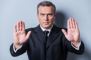 Stop it Serious mature man in formalwear showing his palms and looking at camera while standing against grey background photo