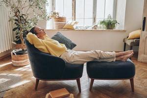 Beautiful young woman holding hands behind head while relaxing in comfortable chair at home photo