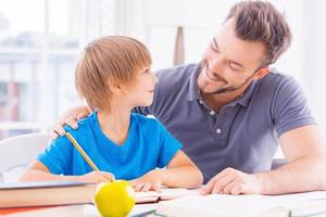 eres un padre joven tan inteligente y alegre que ayuda a su hijo con la tarea mientras se sientan juntos en la mesa foto