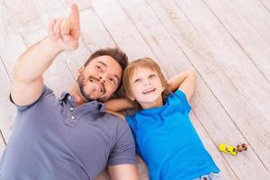 Look over there Top view of happy father and son lying on the hardwood floor together while young man pointing away and smiling photo