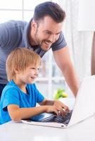 You are my little genius Cheerful father and son looking at the laptop while little boy typing something on keyboard and smiling photo