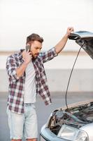 I need help Frustrated young man talking on the mobile phone and holding hand on vehicle hood while standing outdoors photo