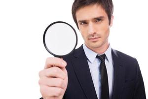 I need details Concentrated young man in formalwear holding magnifying glass and looking at camera while standing isolated on white background photo