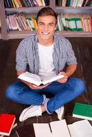 I love studying Top view of happy young man reading book and looking at camera while sitting against bookshelf