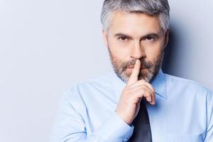 Keep my secret Confident mature man in white shirt and tie looking at camera and holding finger on lips while standing against grey background photo