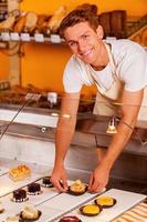 Is this your choice Handsome young man in apron taking a cupcake from showcase and smiling while standing in bakery shop photo