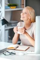 qué hermoso día hermosa mujer joven sosteniendo una taza con bebida caliente y mirando hacia otro lado con una sonrisa mientras se sienta en su lugar de trabajo en la oficina foto