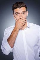 I will keep a secret Surprised young man in white shirt covering mouth with hand and looking at camera while standing against grey background photo