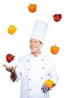 Juggling with colors. Cheerful mature chef in white uniform holding colorful peppers and looking at the red one while standing against white background photo