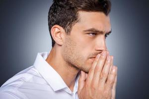 Concentrating at Side view of concentrated young man in white shirt holding hands clasped near face and looking away while standing against grey background photo