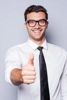 buen trabajo feliz joven con camisa y corbata mostrando su pulgar hacia arriba y sonriendo mientras está de pie contra el fondo gris foto