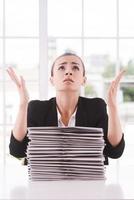 Why me Depressed young woman in suit looking up and gesturing while sitting at the table with stack of documents laying on it photo