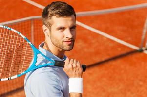 Tennis is my life Top view of happy young man in polo shirt carrying tennis racket on shoulder and smiling while standing on tennis court photo