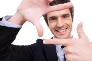 Focus on me Playful young man in formalwear gesturing finger frame and looking through it while standing isolated on white background photo