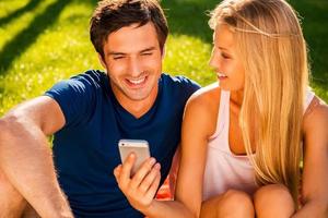 mira a esta feliz pareja joven y amorosa mirando el teléfono móvil y sonriendo mientras se sientan juntos en el césped en el parque foto