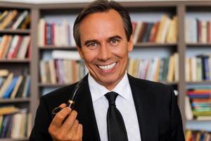 Gentleman with pipe. Portrait of confident mature man in formalwear holding pipe and smiling while standing against bookshelf photo