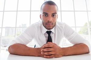 How can I help you Confident young African man in shirt and tie holding hands clasped and looking at camera while sitting at his working place photo