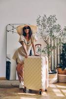 Hermosa mujer joven con sombrero elegante apoyada en la maleta y sonriendo mientras está de pie en casa foto