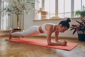 Confident young woman standing in plank position while practicing in front of the window at home photo