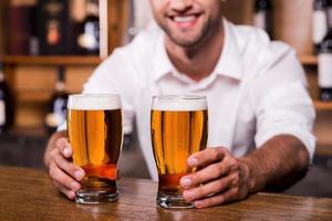 calma tu sed primer plano de un apuesto joven barman con camisa blanca estirando vasos con cerveza y sonriendo mientras está de pie en el mostrador del bar foto