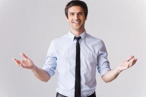 You are always welcome Handsome young man in shirt and tie gesturing and smiling while standing against grey background photo