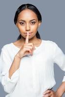 Keep my secret Attractive young African woman holding finger on lips and looking at camera while standing against grey background photo