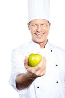 comer sano chef maduro confiado en uniforme blanco estirando manzana verde y sonriendo mientras está de pie contra el fondo blanco foto