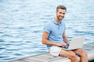 me gusta trabajar al aire libre un joven guapo con camisa de polo trabajando en una laptop y sonriendo mientras estoy sentado en el muelle foto