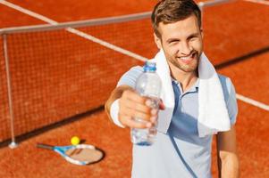 Quench your thirst Happy young man in polo shirt and towel on shoulders stretching out bottle with water while standing on tennis court photo