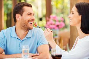 Try my meal Beautiful young woman feeding her boyfriend with salad and smiling while both relaxing in outdoors restaurant photo